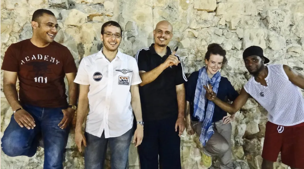 Five diverse people smiling and posing against a stone wall, with one making a peace sign and another pointing upwards.