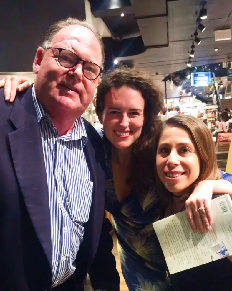 Three people smiling indoors, with a man wearing glasses and a striped shirt on the left, and two women on the right, one holding a book.
