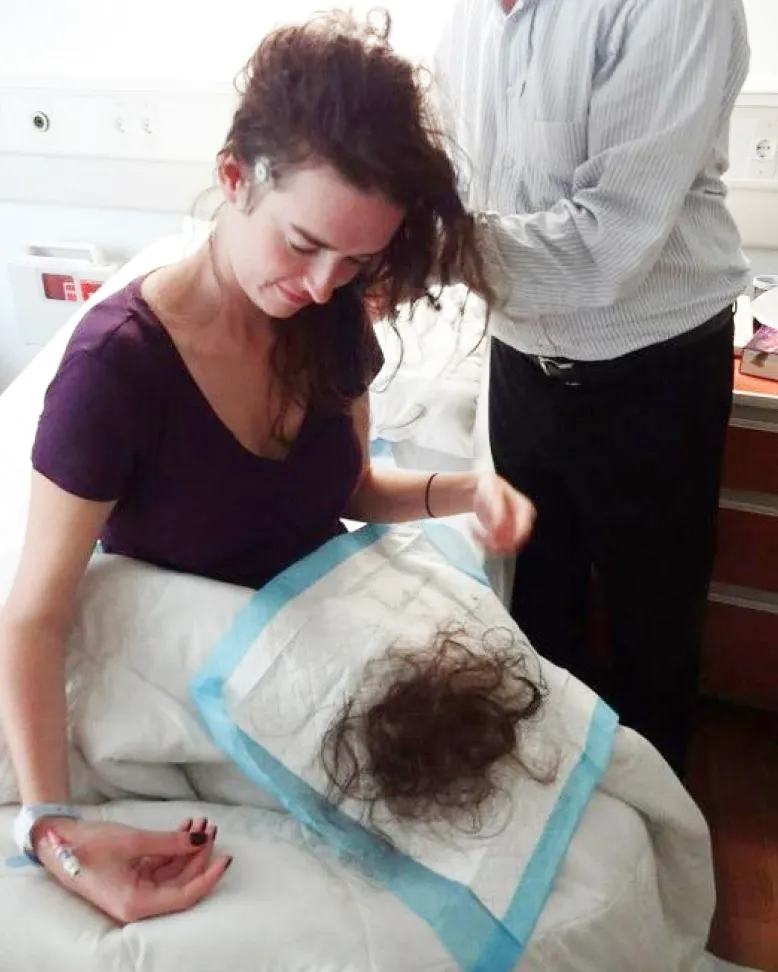 Woman in hospital bed looking down at cut hair on pad while caregiver trims her hair in preparation for a medical procedure