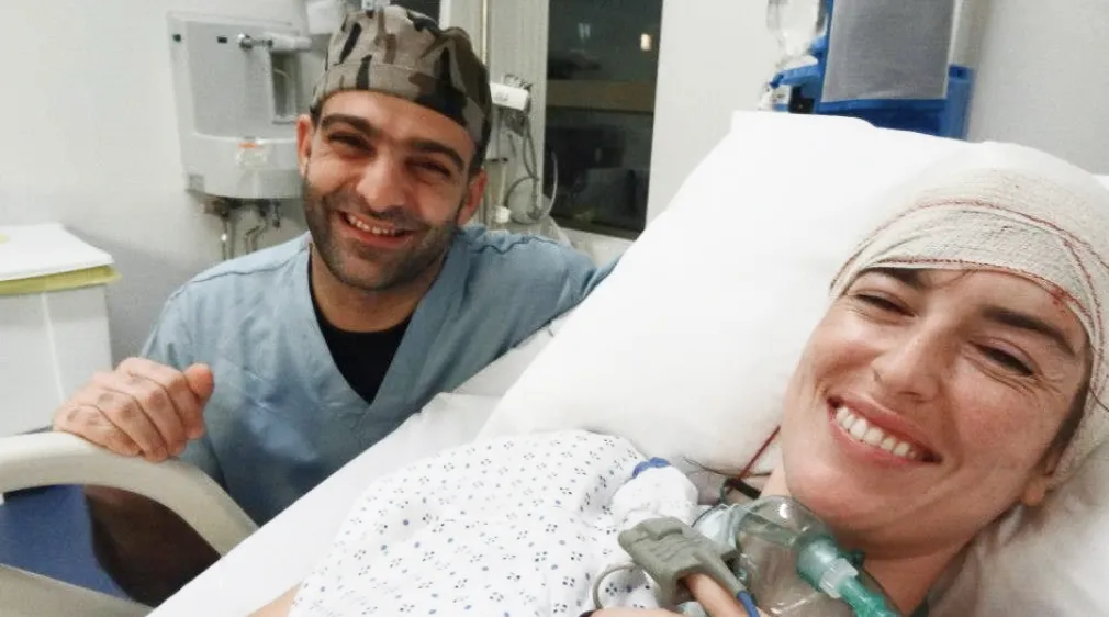 Smiling woman in hospital bed with head bandage holding oxygen mask, next to supportive man in medical scrubs