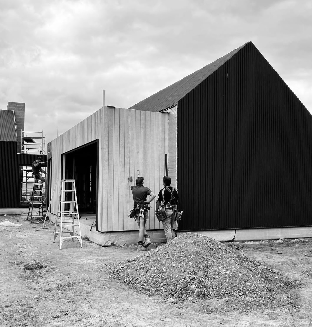Two construction workers discussing near a wooden building under construction, with ladders and scaffolding visible.