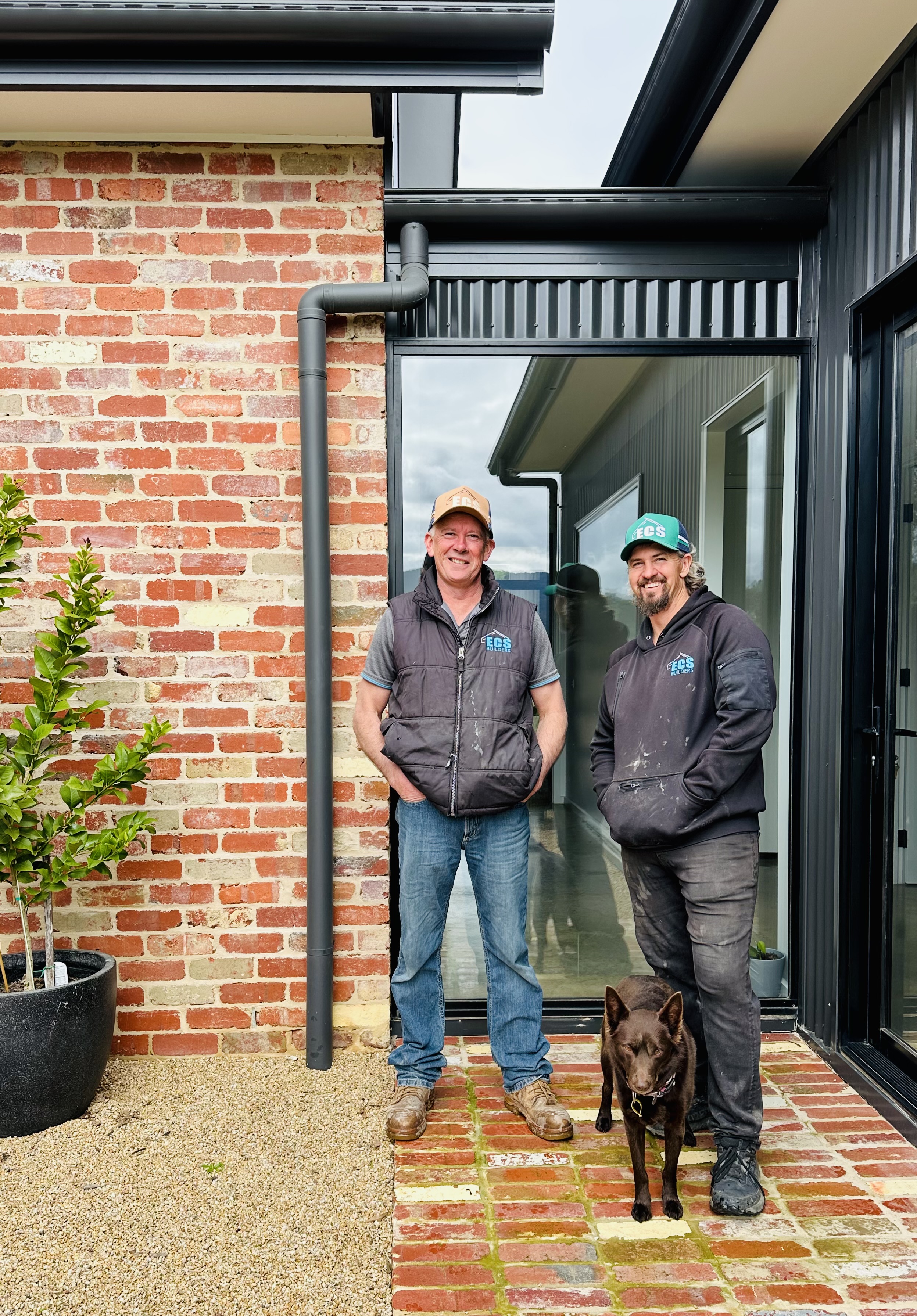 Two men wearing ECS Builders hats and jackets standing on a brick patio outside a modern house, with a brown dog beside them.