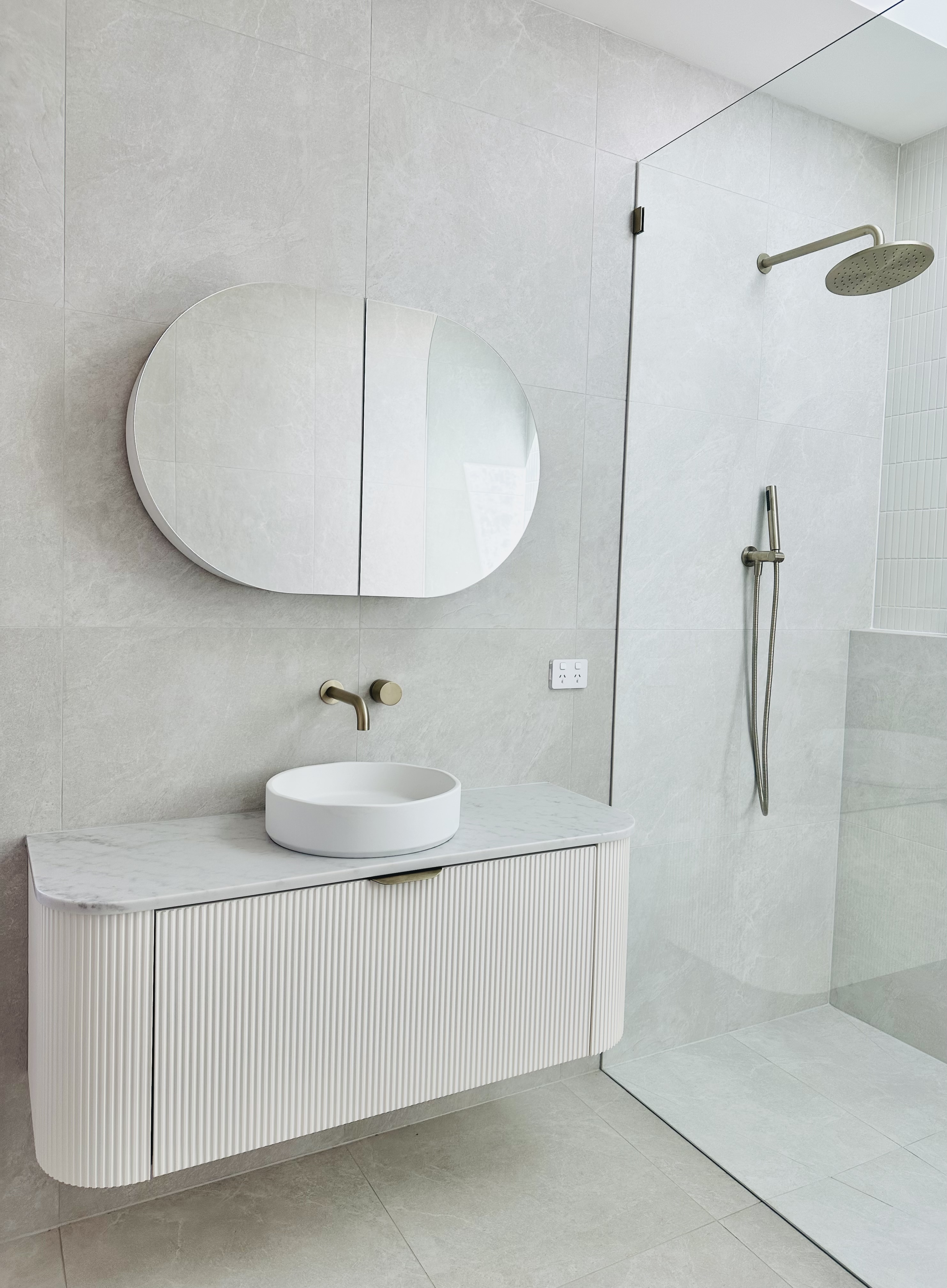 Minimalist bathroom with a wall-mounted floating vanity with a round vessel sink, a large oval mirror, and a glass-enclosed shower with a rainfall showerhead.
