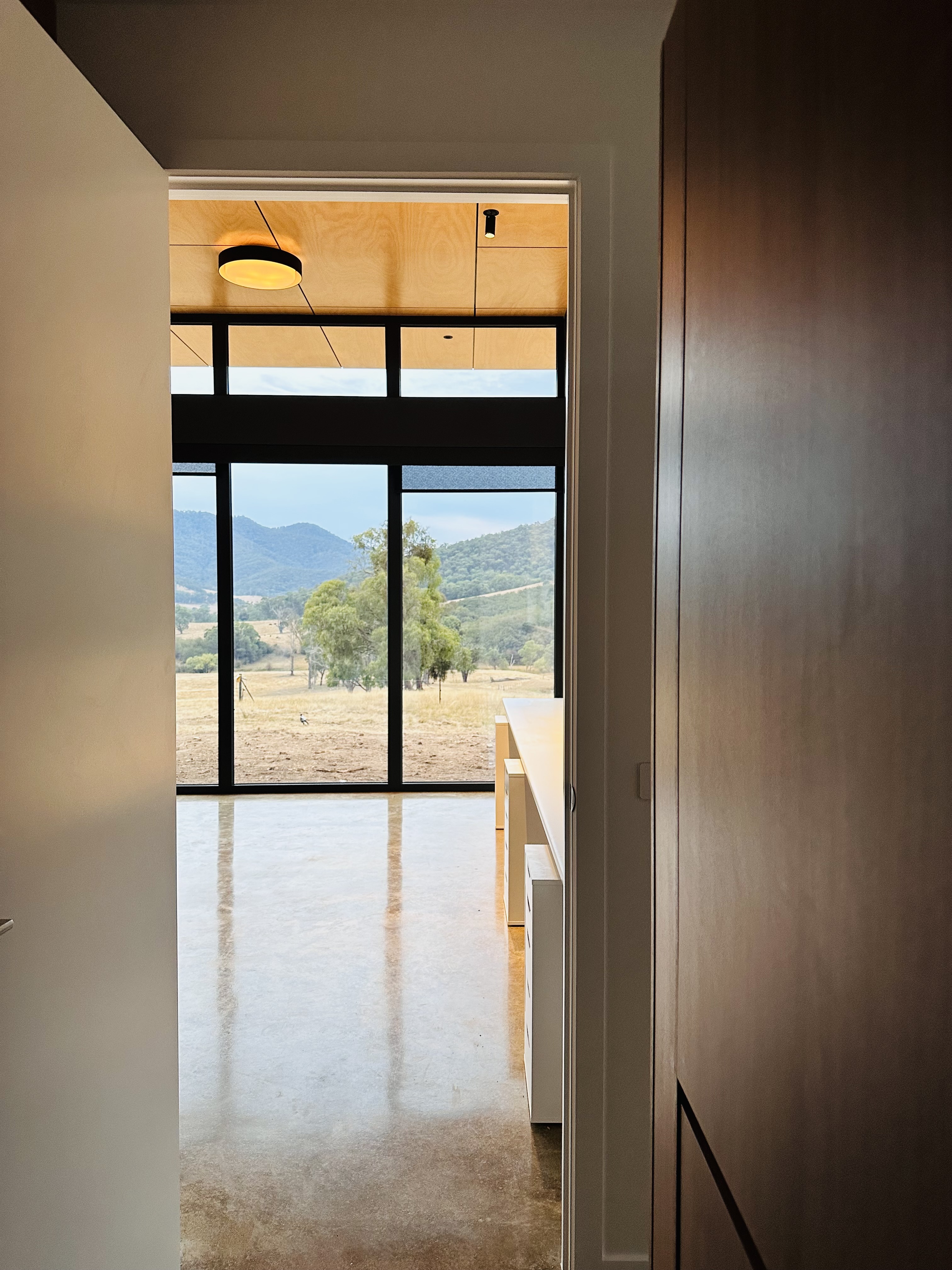 Interior view through an open door showing a room with polished concrete floors, large windows, and a landscape of trees and hills outside.