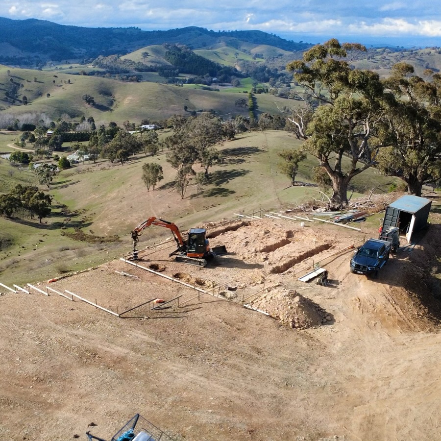 Excavator digging at a construction site on a hill with rolling green hills and trees in the background.