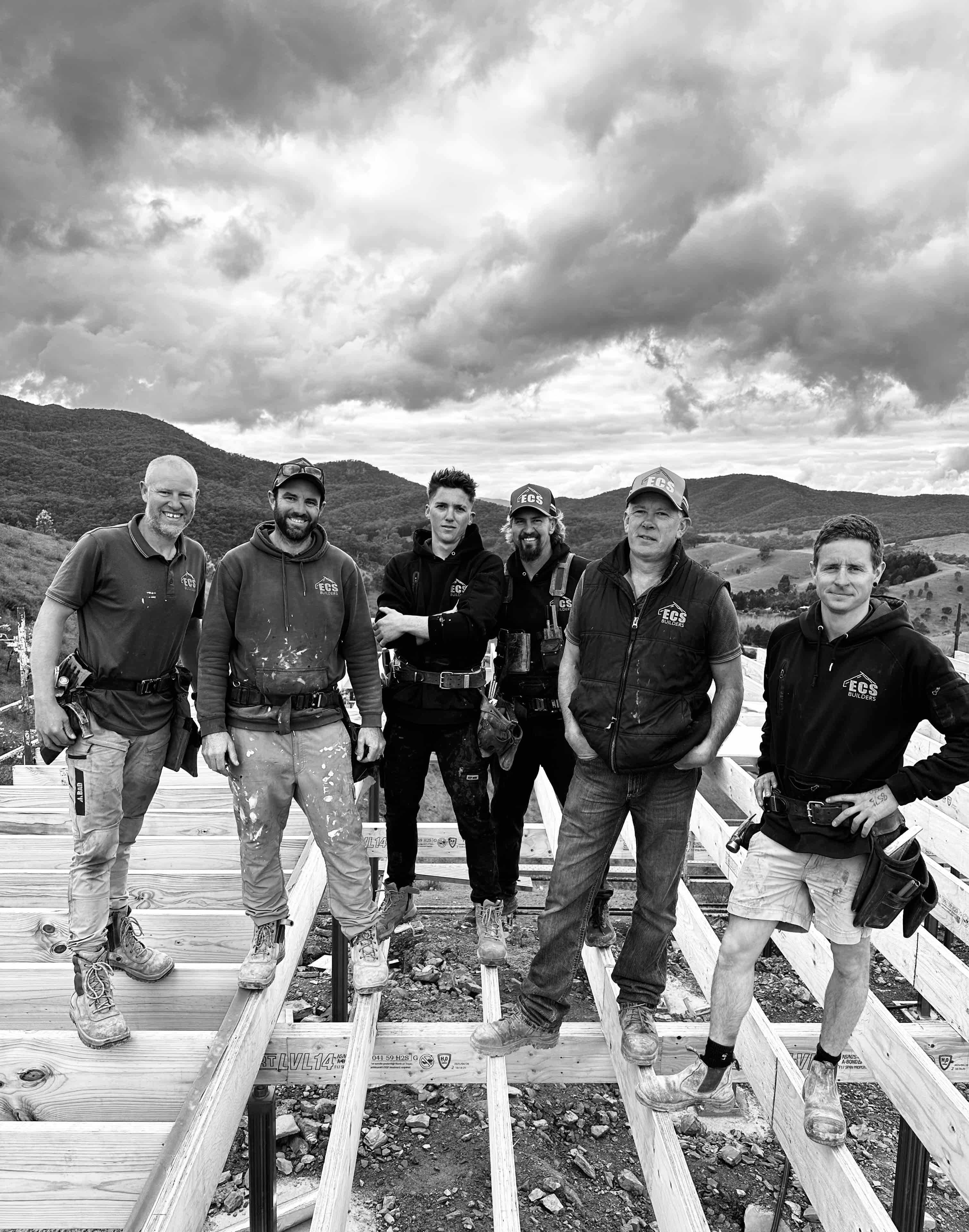Six construction workers standing on wooden beams at a building site with hills and cloudy sky in the background.