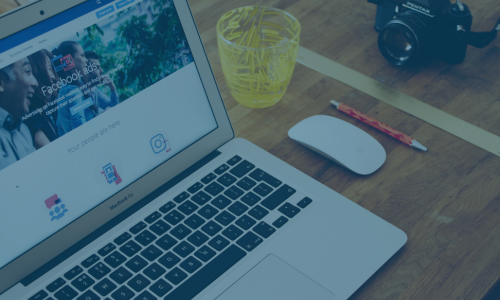 Laptop on a wooden desk displaying a Facebook Ads page with a yellow pencil cup, white mouse, and a camera nearby.