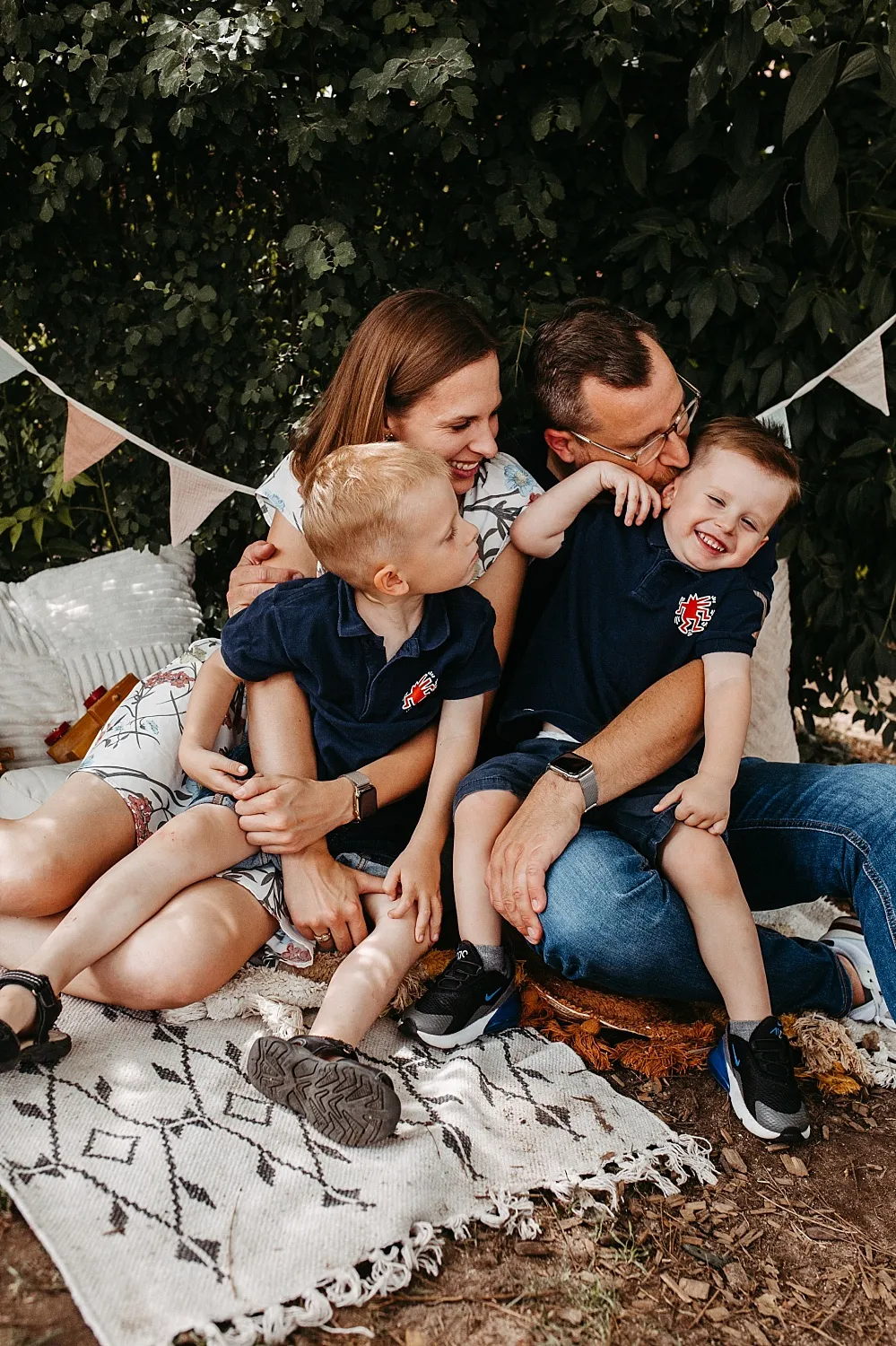 Elternduo sitzt draußen auf einer Decke und umarmt lachende zwei Jungen in blauem Poloshirt.