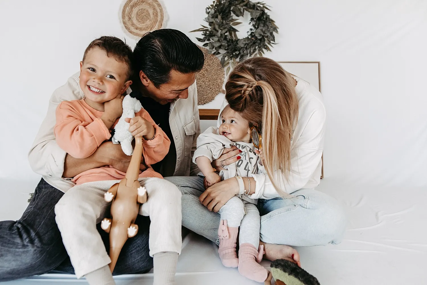 Familie mit zwei Kindern sitzt auf dem Boden, die Eltern lächeln und umarmen ihre Kinder spielerisch.