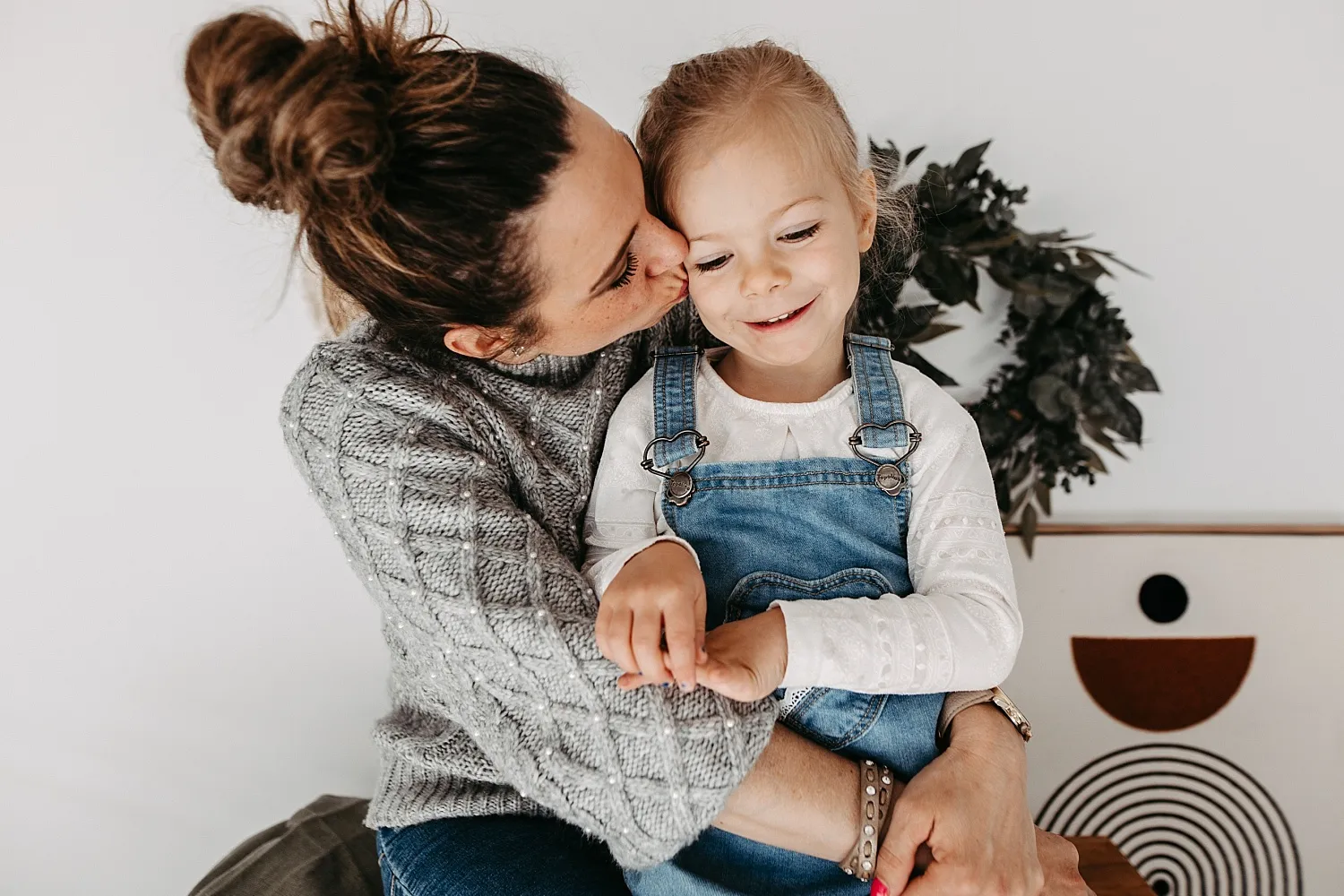 Eine Frau im grauen Pullover umarmt und küsst liebevoll ein lächelndes kleines Mädchen in blauer Latzhose.