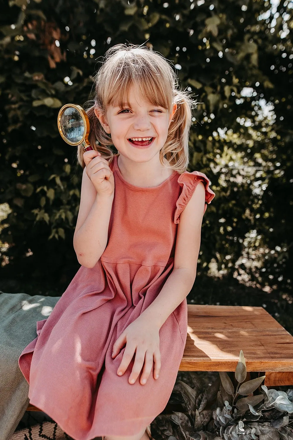 Lachendes Mädchen mit Zöpfen, das eine Lupe hält, sitzt auf einer Holzbank im Freien.