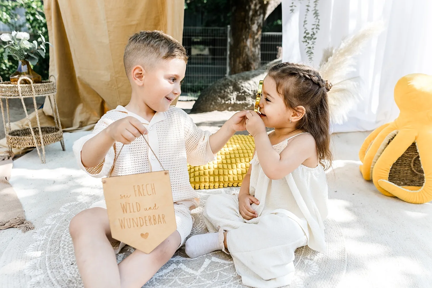 Zwei Kinder sitzen auf einem Teppich, der Junge hält ein Schild mit der Aufschrift „sei frech wild und wunderbar“, während das Mädchen durch eine Lupe schaut.