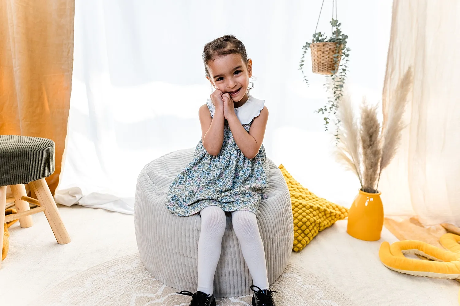 Ein lächelndes kleines Mädchen sitzt auf einem grauen gepolsterten Sitzhocker vor weißem Vorhang mit gelben Kissen und Dekoration.