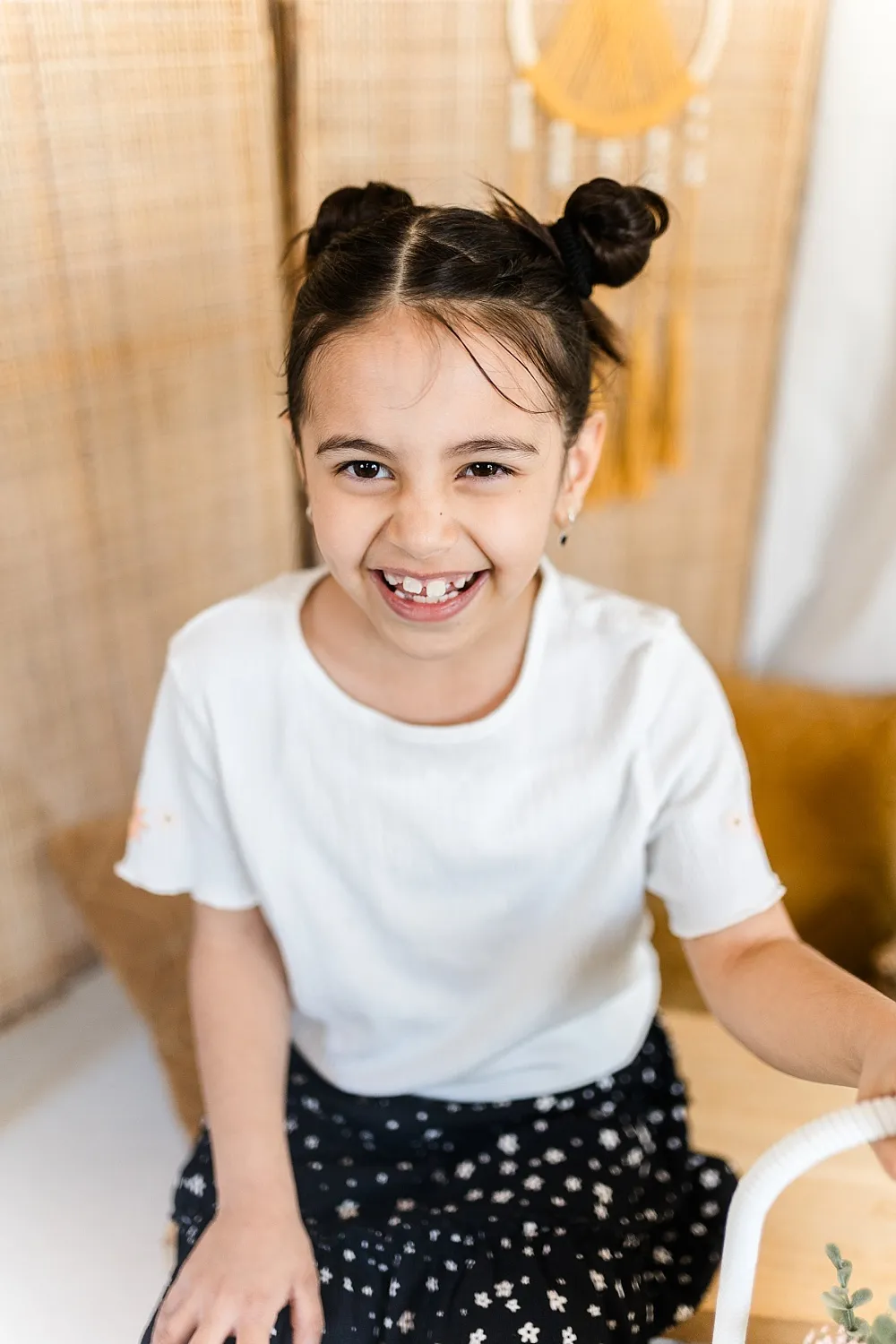 Lachendes Mädchen mit weißen Zähnen und zwei Haarknoten trägt ein weißes T-Shirt und einen schwarzen Rock mit weißen Blumen.