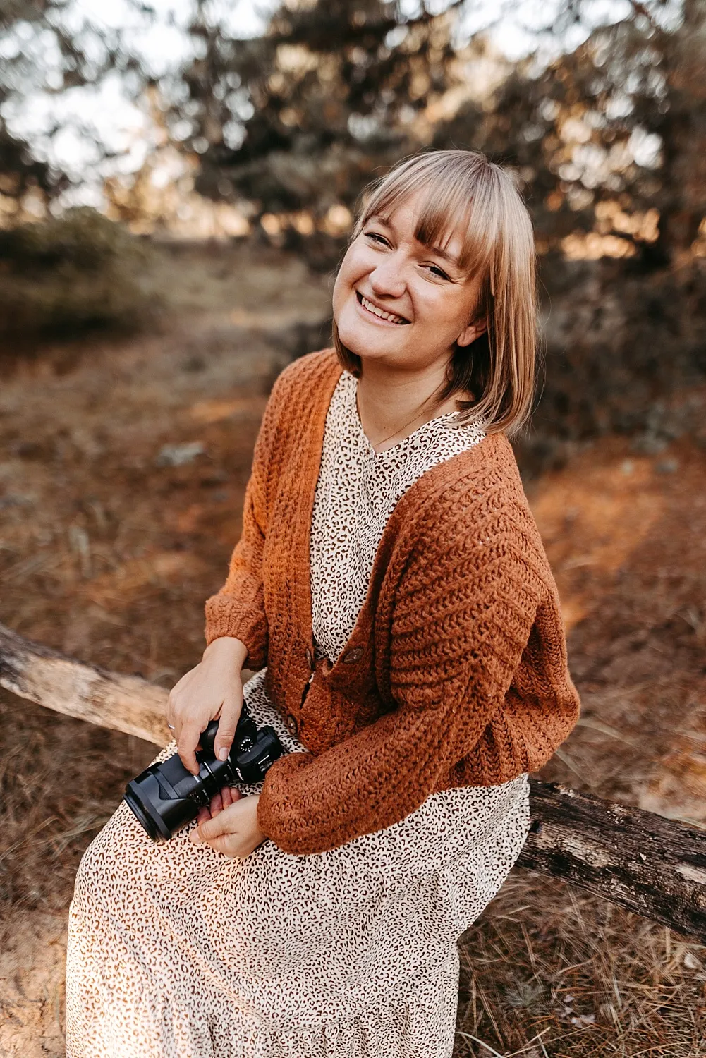 Lächelnde Frau mit kurzem blonden Haar in braunem Strickcardigan und gemustertem Kleid sitzt im Wald auf einem Baumstamm und hält eine Kamera.