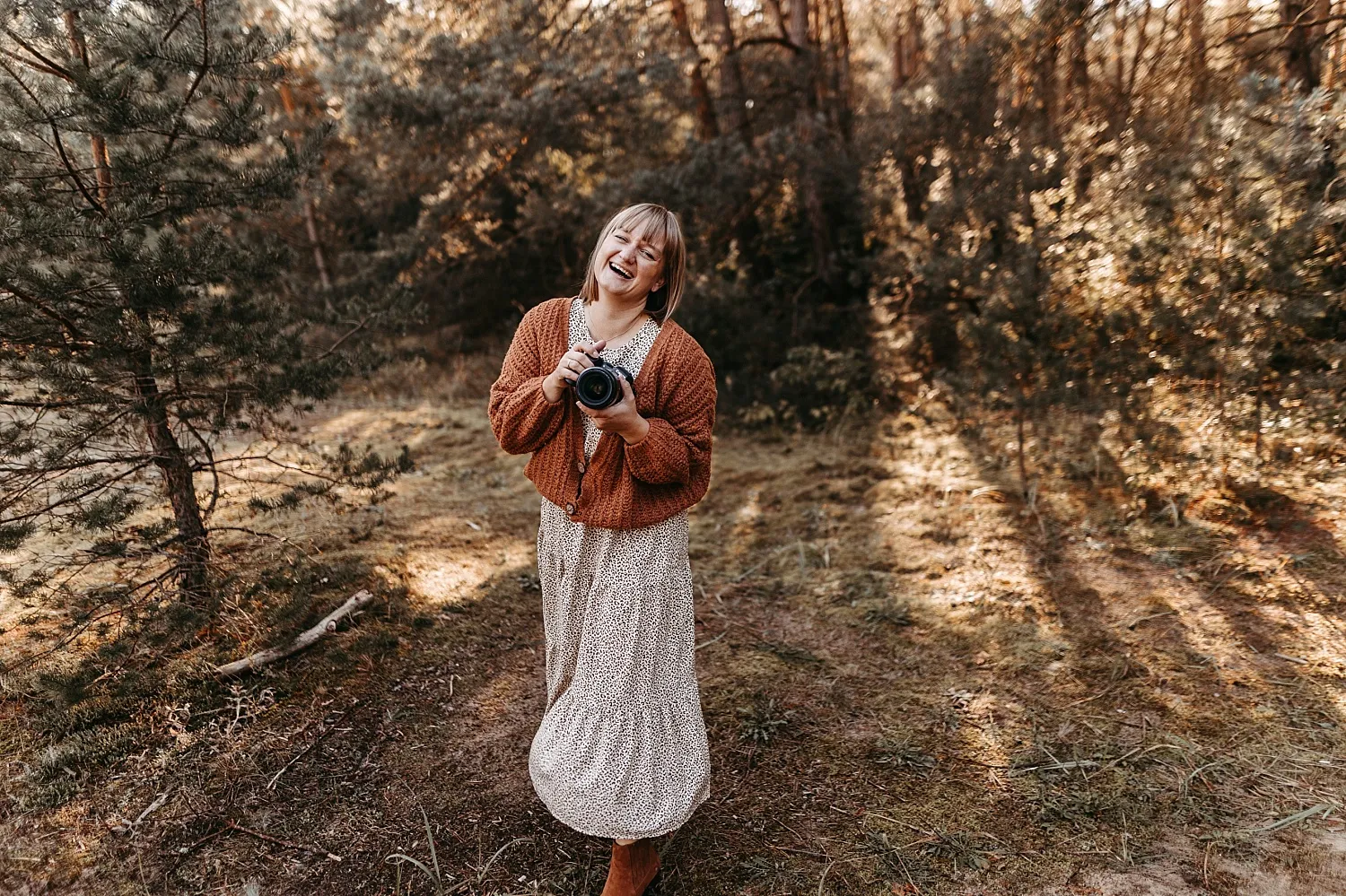 Lächelnde Frau mit brauner Strickjacke und Kleid hält eine Kamera in einem sonnenbeschienenen Wald.