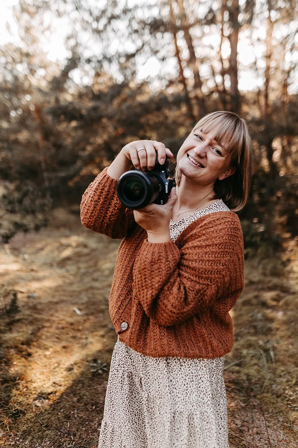 Frau mit kurzem blonden Haar und karierter Kleid trägt eine braune Strickjacke und hält eine Kamera im Wald.