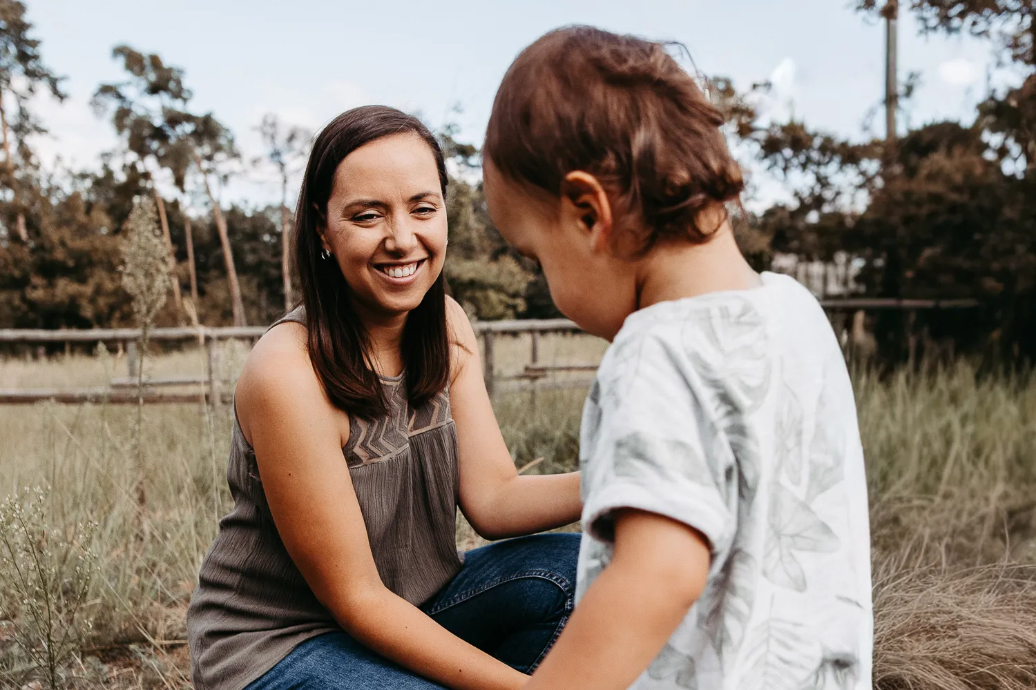 Lächende Frau sitzt im Freien auf dem Boden und spielt mit einem kleinen Jungen, umgeben von Gras und Bäumen.