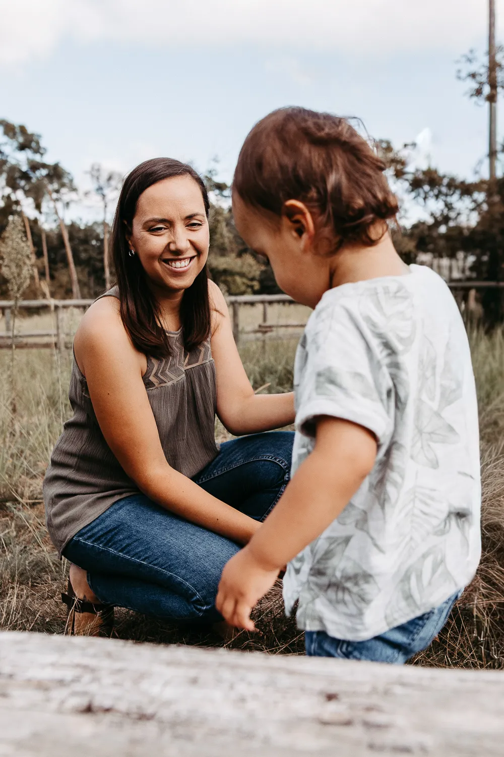 Frau kauert lächelnd im Gras und schaut auf Kleinkind in weißem T-Shirt und Jeans.