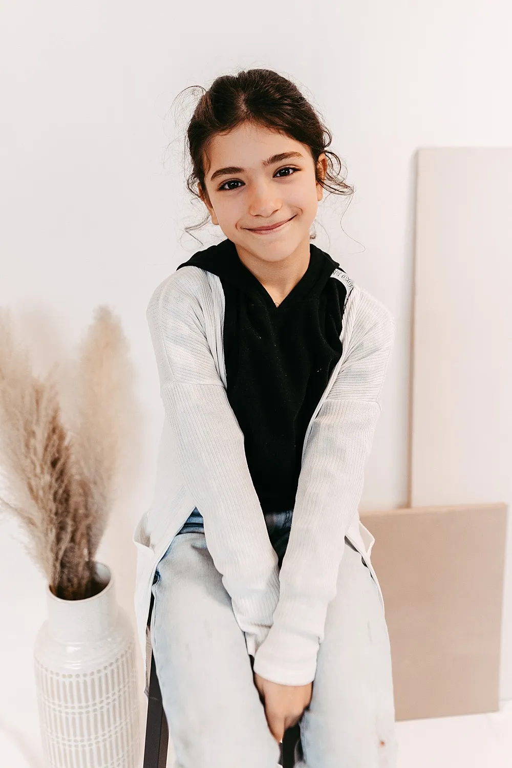Lächeldes Mädchen mit dunklen Haaren trägt schwarzen Pullover und weiße Strickjacke, sitzt vor hellem Hintergrund mit Vase und getrocknetem Pampasgras.