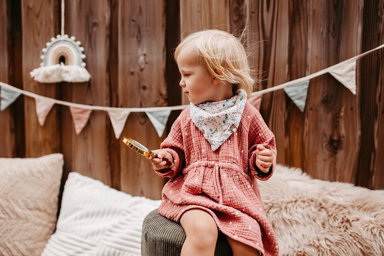 Kleines blondes Mädchen in rosa Kleid mit Halstuch hält eine Lupe und sitzt vor einer Holzpaneelwand mit Wimpelgirlande.