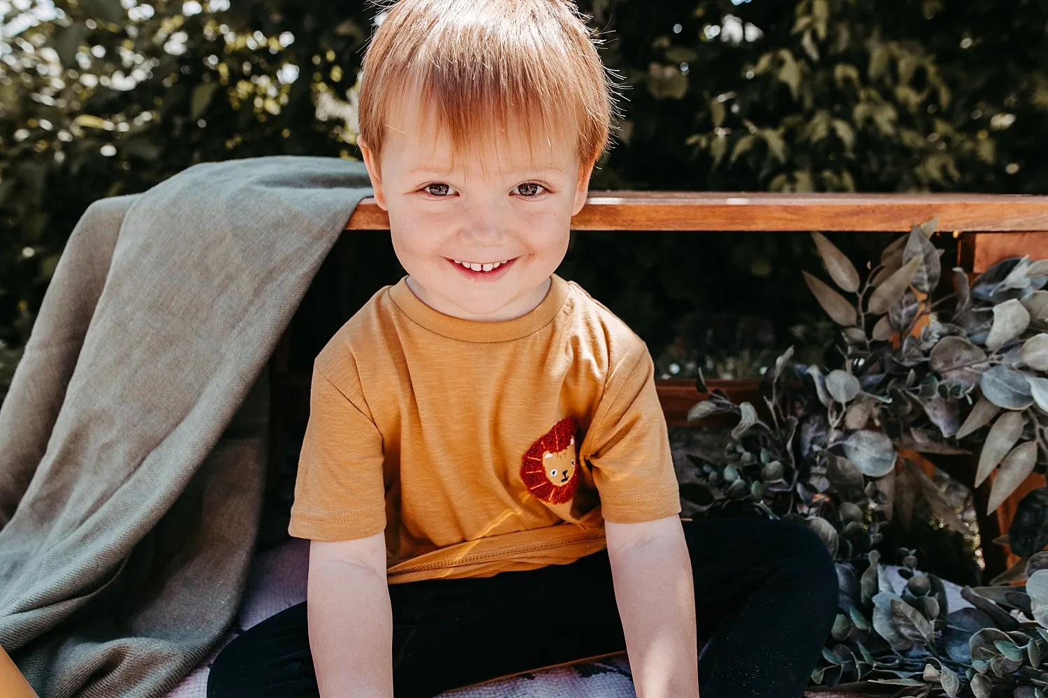 Lächelnder kleiner Junge mit orangefarbenem T-Shirt und Löwenmotiv sitzt auf einer Decke vor grünem Laub.