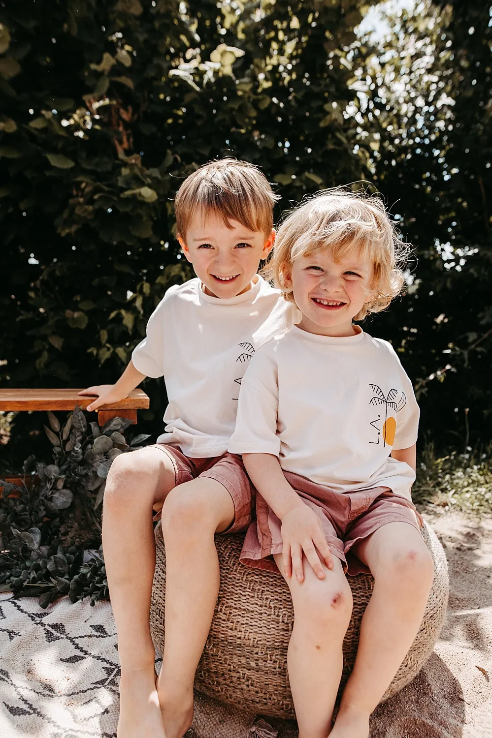 Zwei lächelnde kleine Jungen sitzen draußen auf einem runden Häkelkissen, tragen weiße T-Shirts und rosa Shorts.