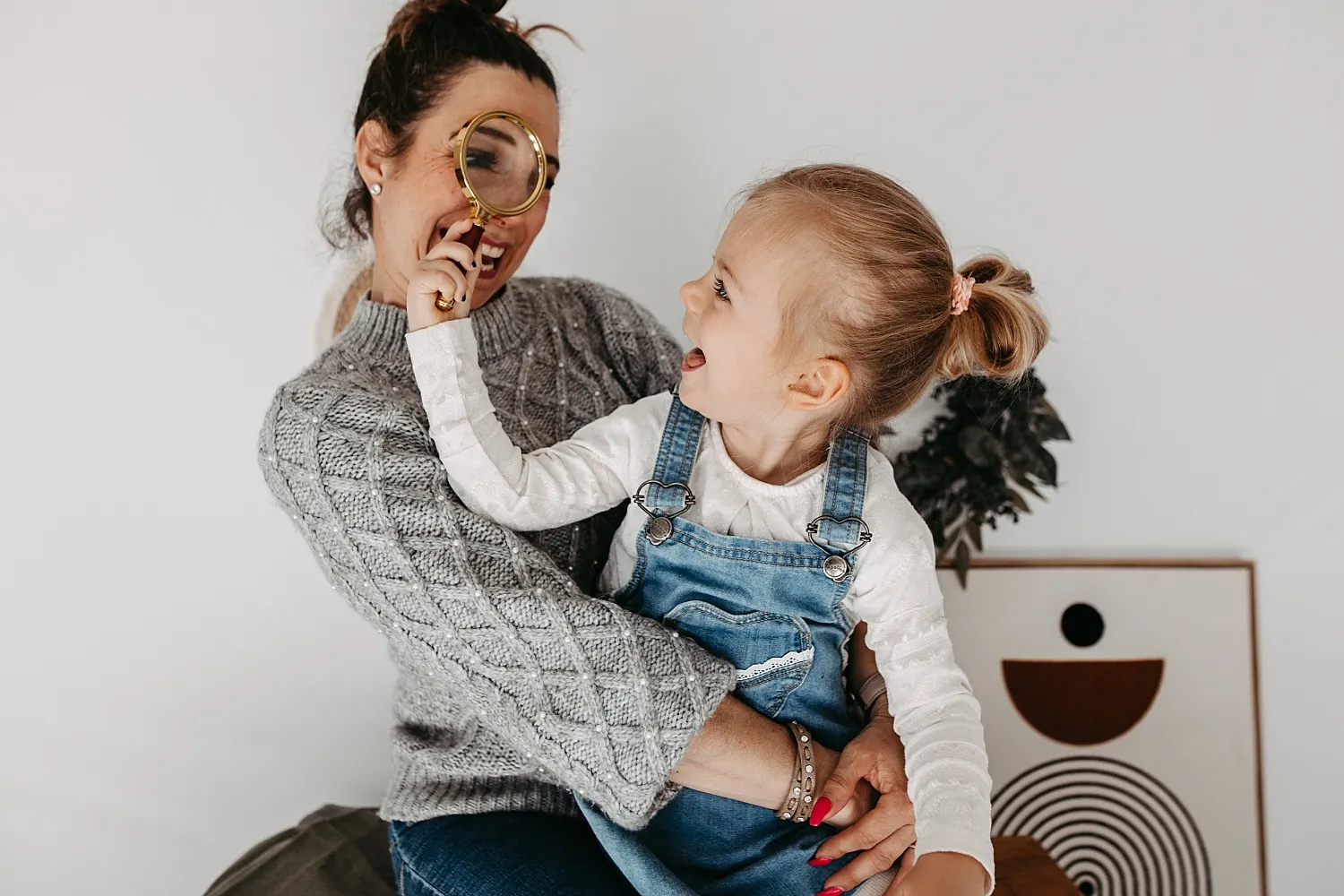 Ein kleines Mädchen hält eine Lupe vor das Gesicht einer lächelnden Frau, die sie auf dem Schoß hält.