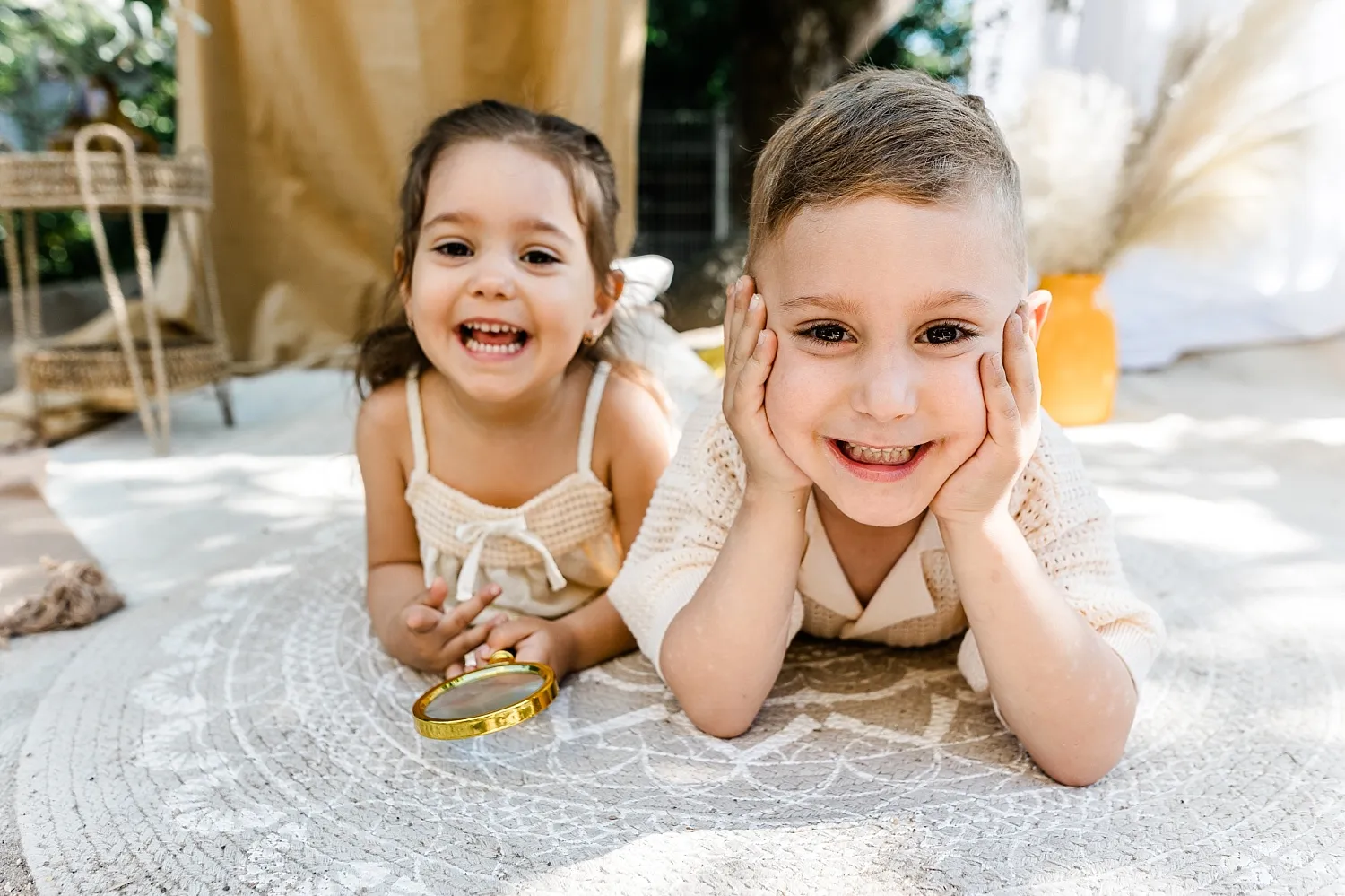Zwei lächelnde Kinder liegen auf einem Teppich, das Mädchen hält eine Lupe.