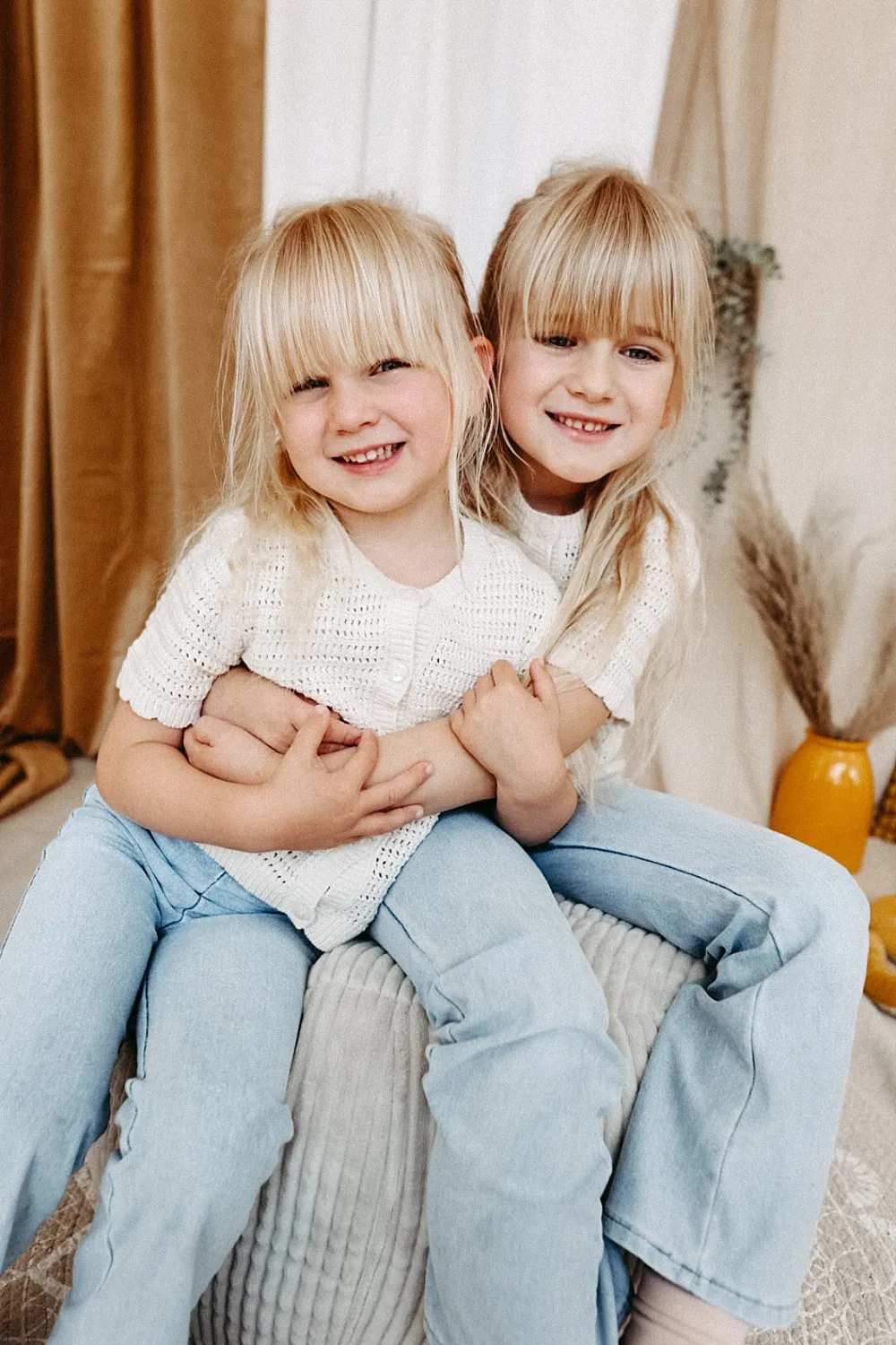 Zwei blonde Mädchen in weißen Pullovern und blauen Jeans umarmen sich lachend auf einem weichen Sitz.