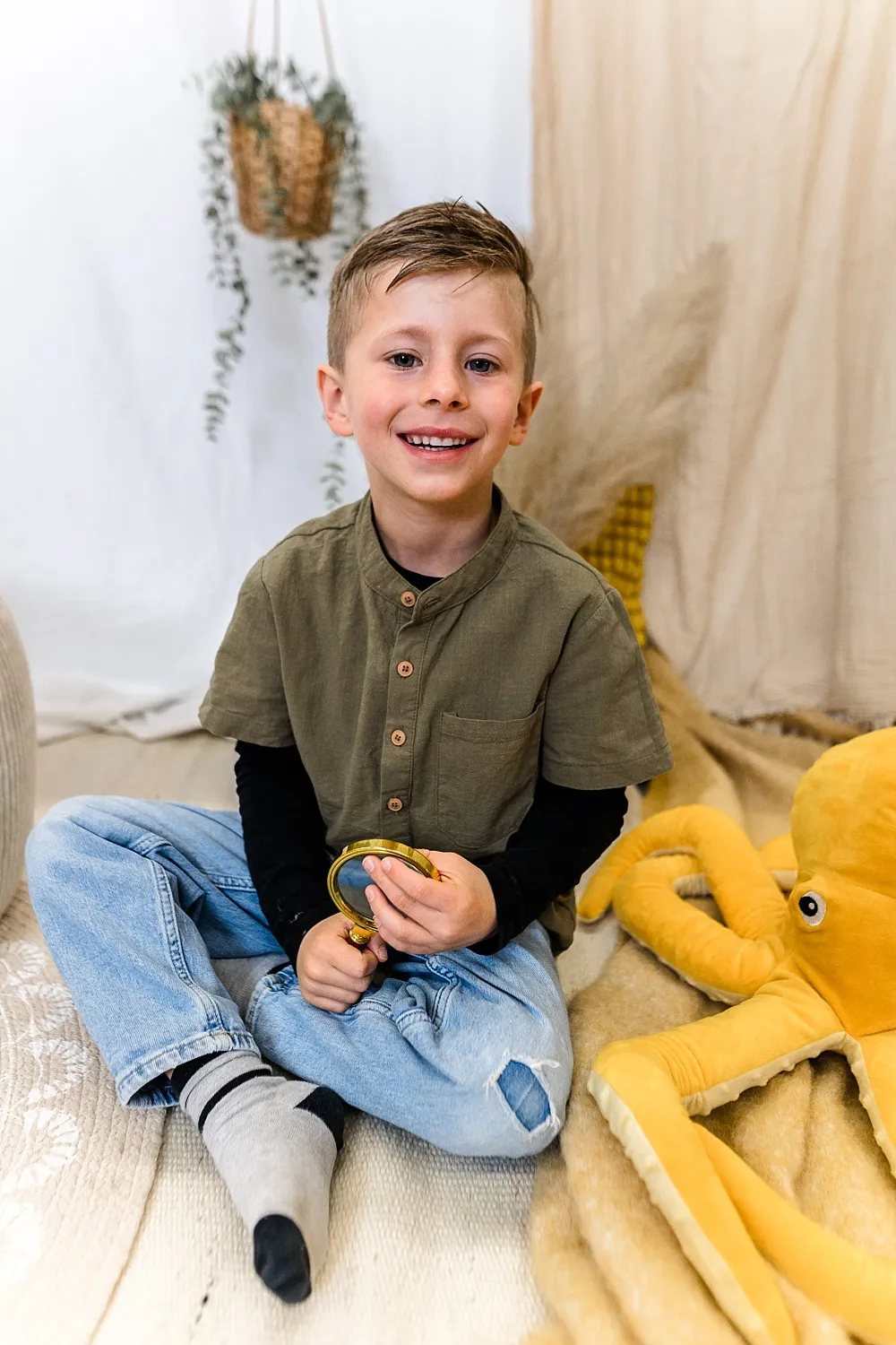 Lächelnder Junge sitzt mit Lupe in der Hand auf dem Boden neben einem großen gelben Stoff-Oktopus.