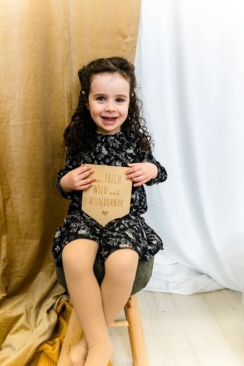 Lächendes Mädchen mit lockigem Haar, das ein Schild mit der Aufschrift ‚frech wild und wunderbar‘ hält, sitzt auf einem Hocker vor braunem und weißem Vorhang.