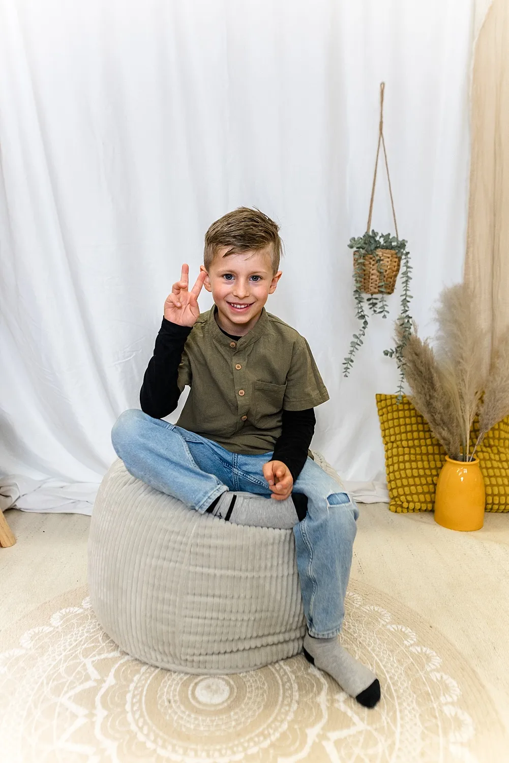 Ein Junge sitzt verschränkt auf einem grauen Sitzpouf und zeigt mit der rechten Hand ein Peace-Zeichen, im Hintergrund hängen Pflanzen und Kissen.
