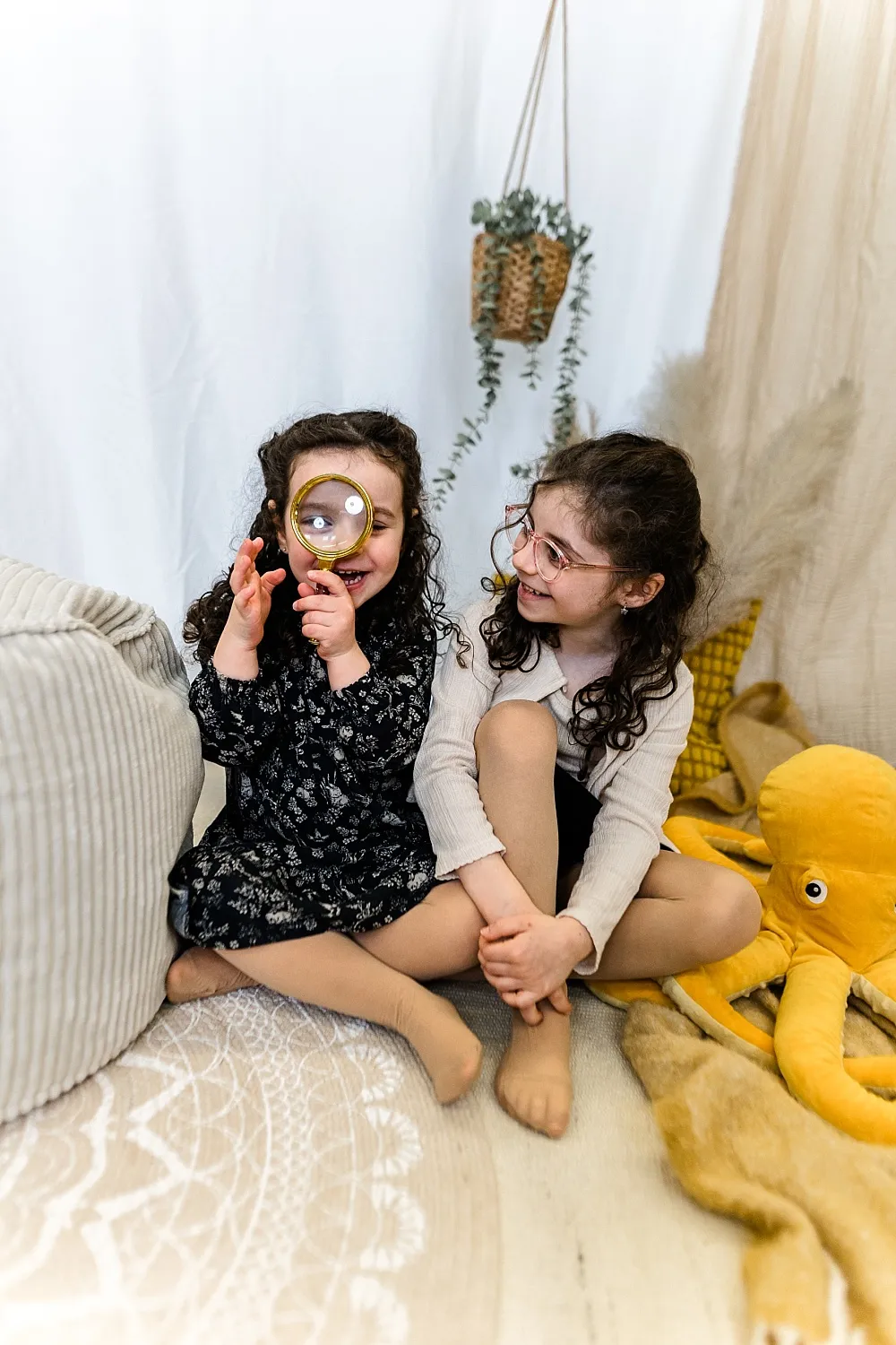 Zwei lächelnde Mädchen mit lockigem Haar sitzen auf einem Teppich, eines hält eine Lupe vor ein Auge, neben ihnen liegt ein großes gelbes Tintenfisch-Kuscheltier.