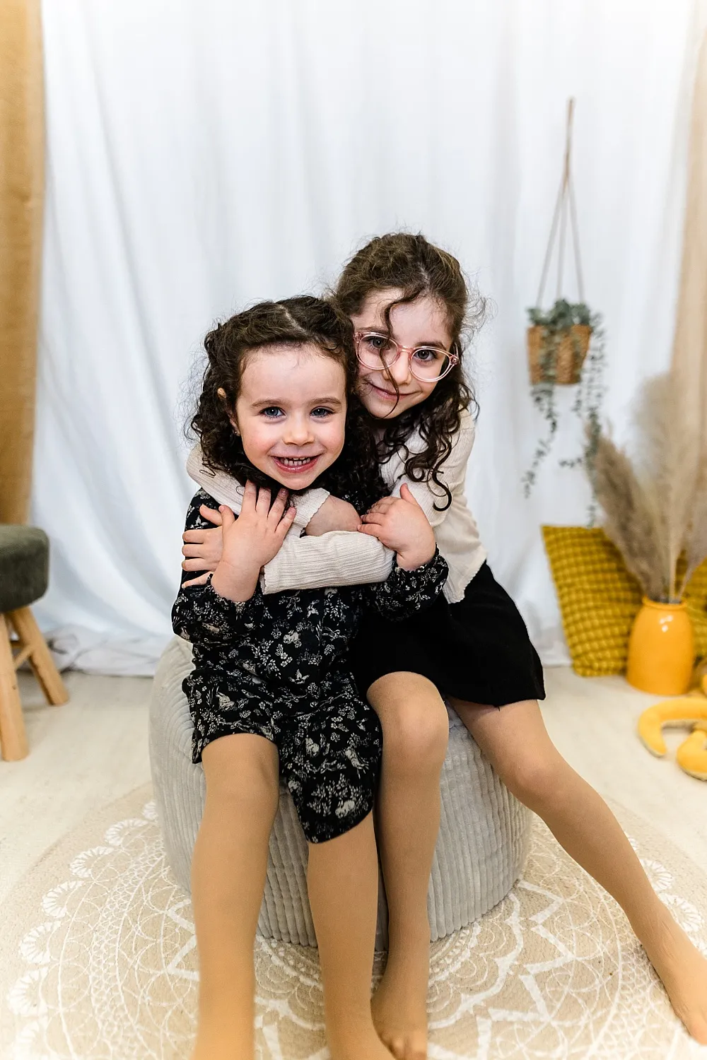 Zwei junge Mädchen mit lockigem Haar sitzen lachend und umarmen sich auf einem grauen Sitzpouf in einem hellen Raum mit gelben Dekokissen und Pflanzen.