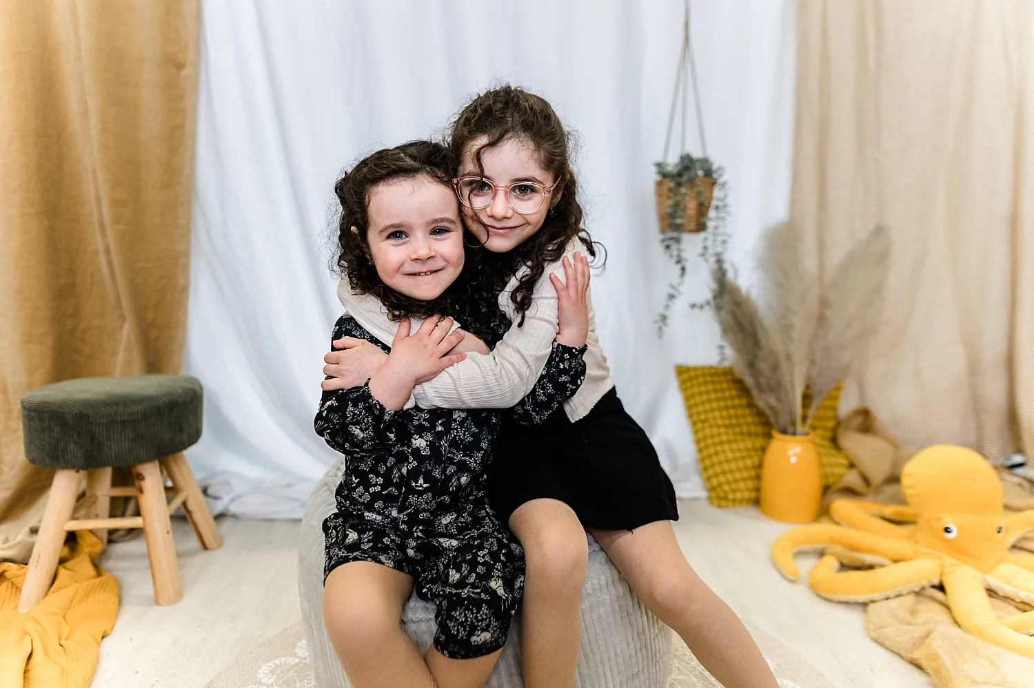 Zwei Mädchen mit lockigem Haar umarmen sich lächelnd in einem hellen Raum mit gemütlicher Dekoration.