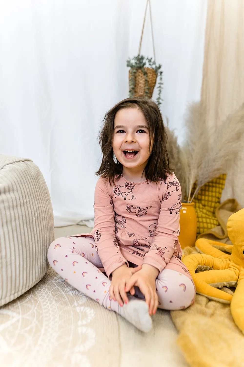 Lachendes Mädchen mit rosa Einhornpyjama sitzt auf dem Boden in einem gemütlichen, hellen Raum.