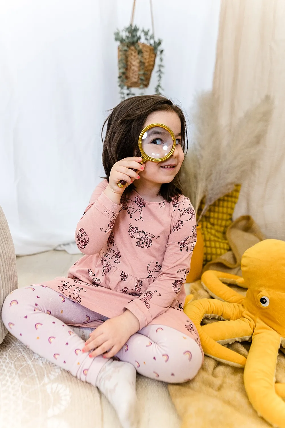 Mädchen sitzt auf dem Boden mit rosa Kleid und hält eine Lupe vor einem Auge, neben ihr ein großes gelbes Plüsch-Oktopus-Spielzeug.