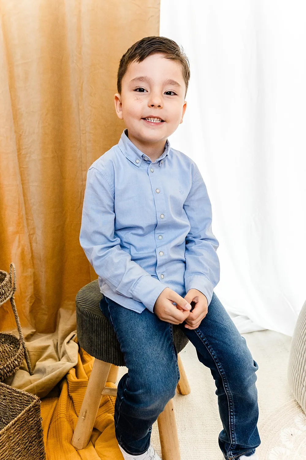 Junge mit blauem Hemd und Jeans sitzt lächelnd auf einem Hocker vor braunem Vorhang und weißem Hintergrund.