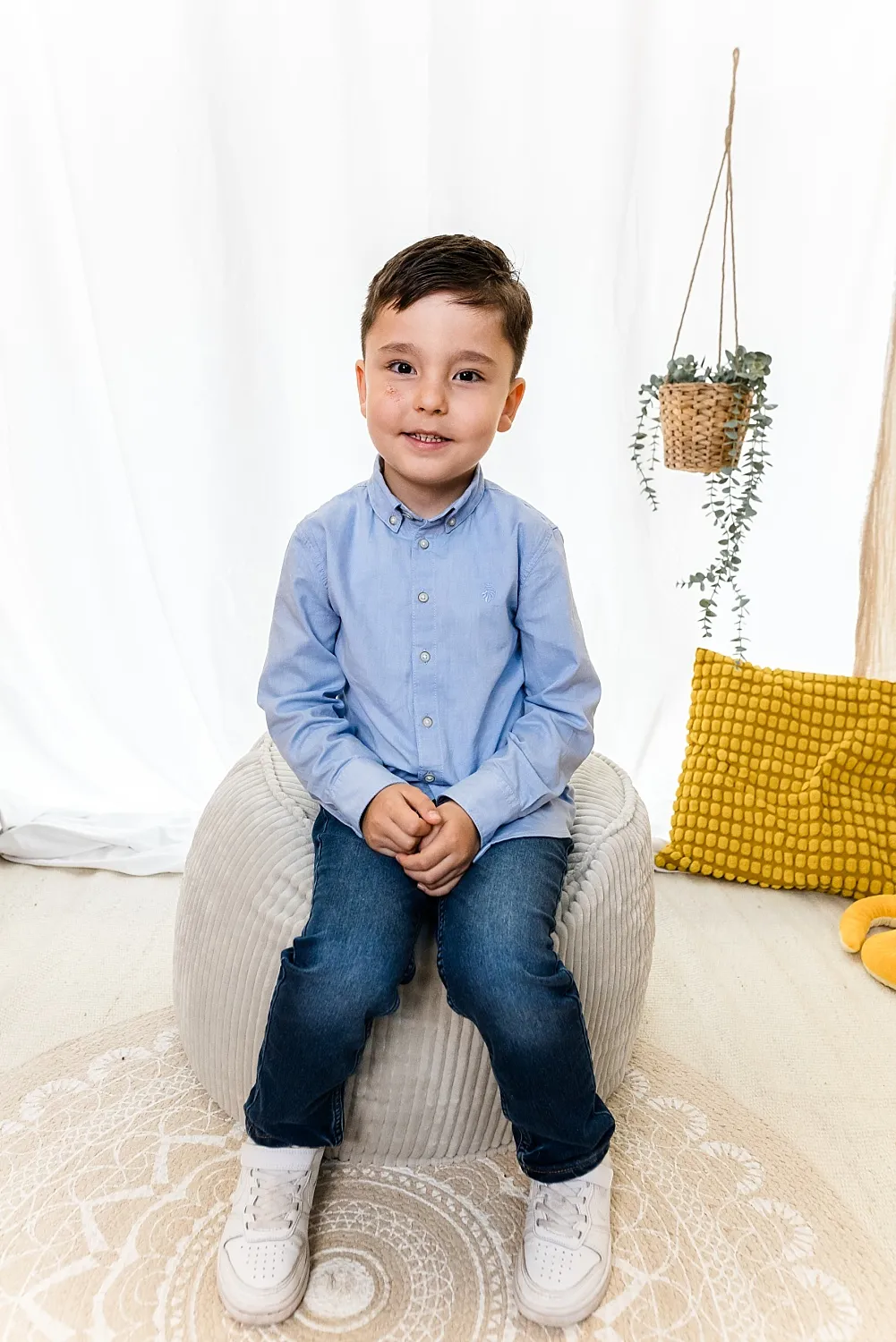 Junge mit blauem Hemd und Jeans sitzt auf einem grauen Sitzpouf vor weißem Hintergrund mit gelbem Kissen und hängender Pflanze.