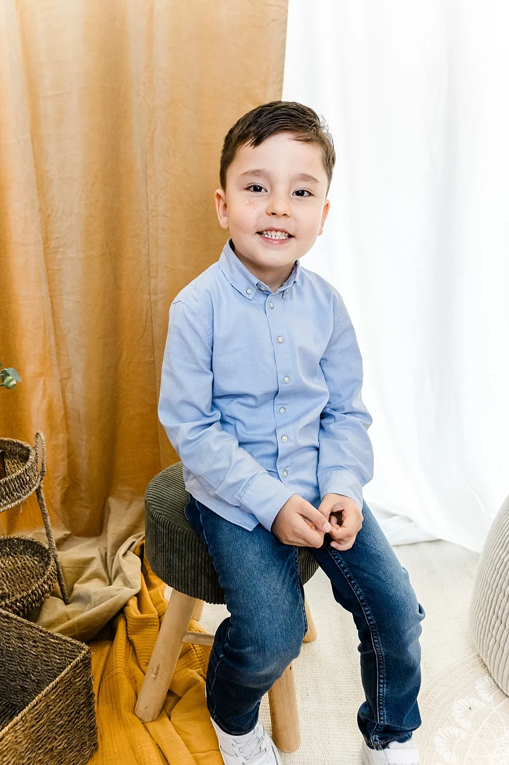 Kleiner Junge mit hellblauem Hemd und Jeans sitzt lächelnd auf einem Hocker vor beigen und weißen Vorhängen.