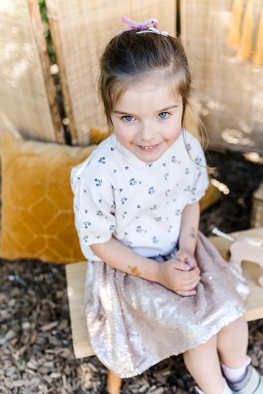 Ein lächelndes Mädchen mit blauen Augen sitzt auf einer Holzbank, trägt ein weißes Oberteil mit Blumenmuster und einen hellen, glitzernden Rock.