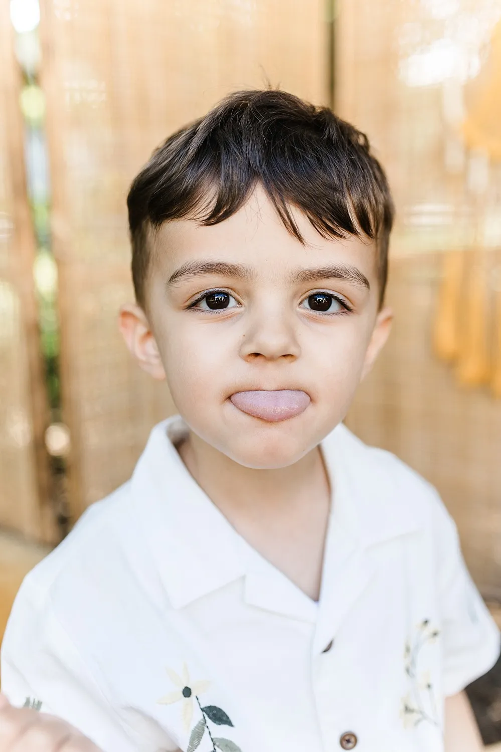 Kleiner Junge mit dunklen Haaren streckt die Zunge heraus und trägt ein weißes Hemd mit Blumendruck.