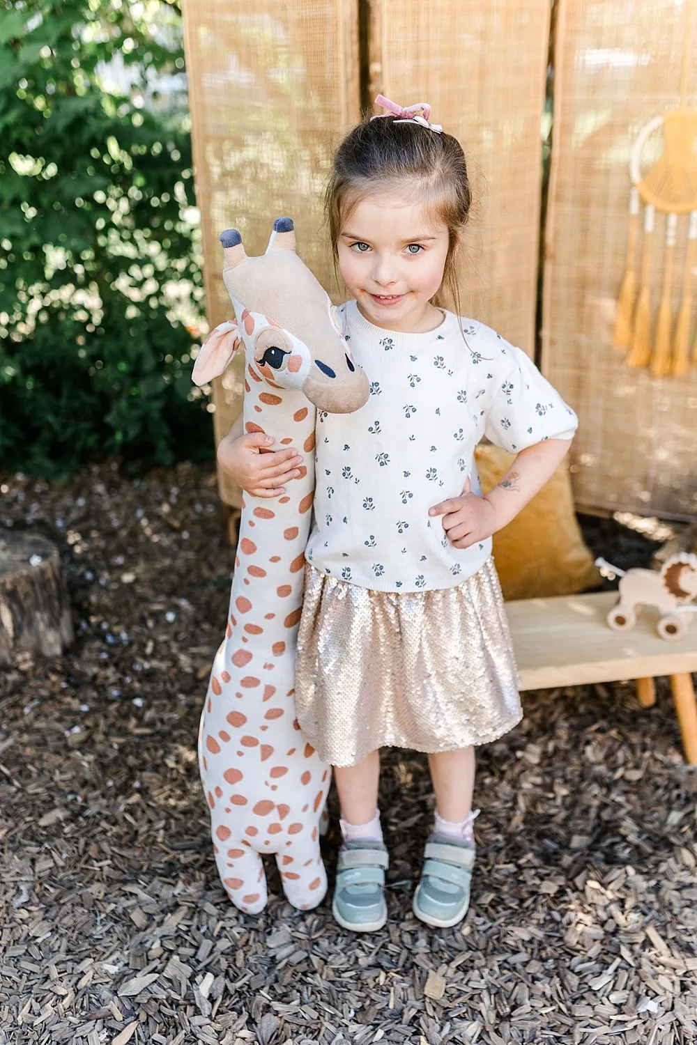 Kleines Mädchen mit hellem Pullover und Rock steht draußen und hält ein großes Plüschgiraffen-Spielzeug.