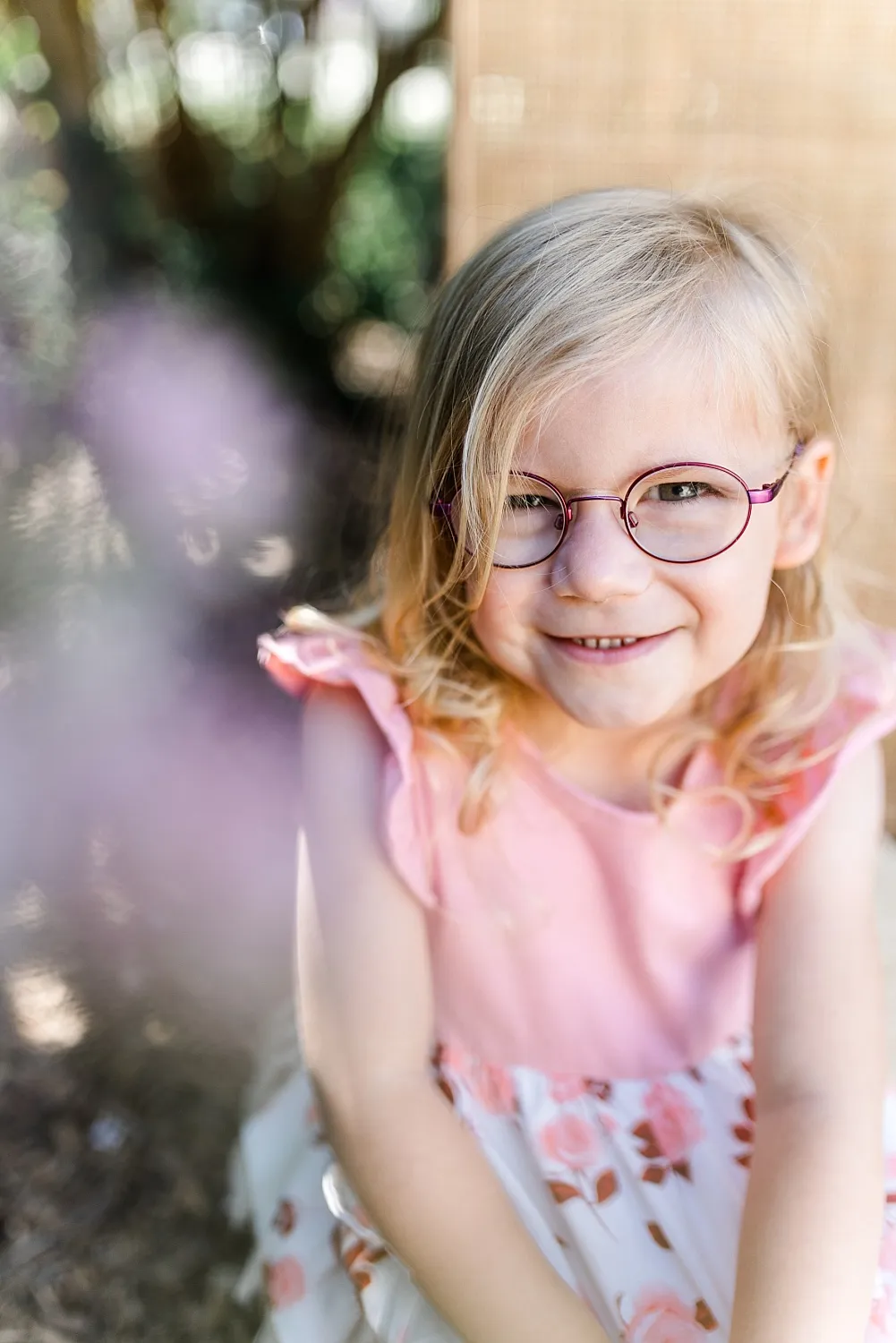 Lächeldes blondes Mädchen mit Brille in rosa Kleid mit Blumenmuster vor verschwommenem Naturhintergrund.