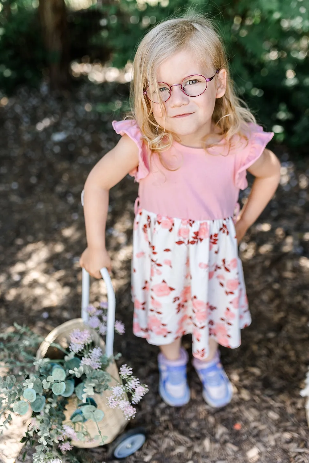 Kleines Mädchen mit Brille in einem rosa-weißen Blumenkleid, das einen Wagen mit Pflanzen zieht.