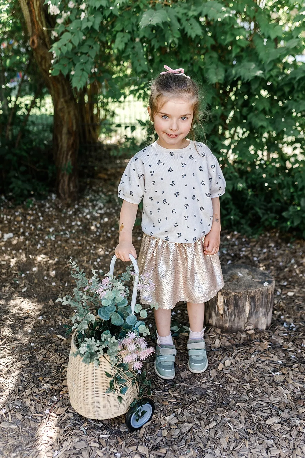 Kleines Mädchen mit rosa Haarspange und goldfarbenem Rock steht im Garten und zieht einen Korbwagen mit grünen und rosa Blumen.