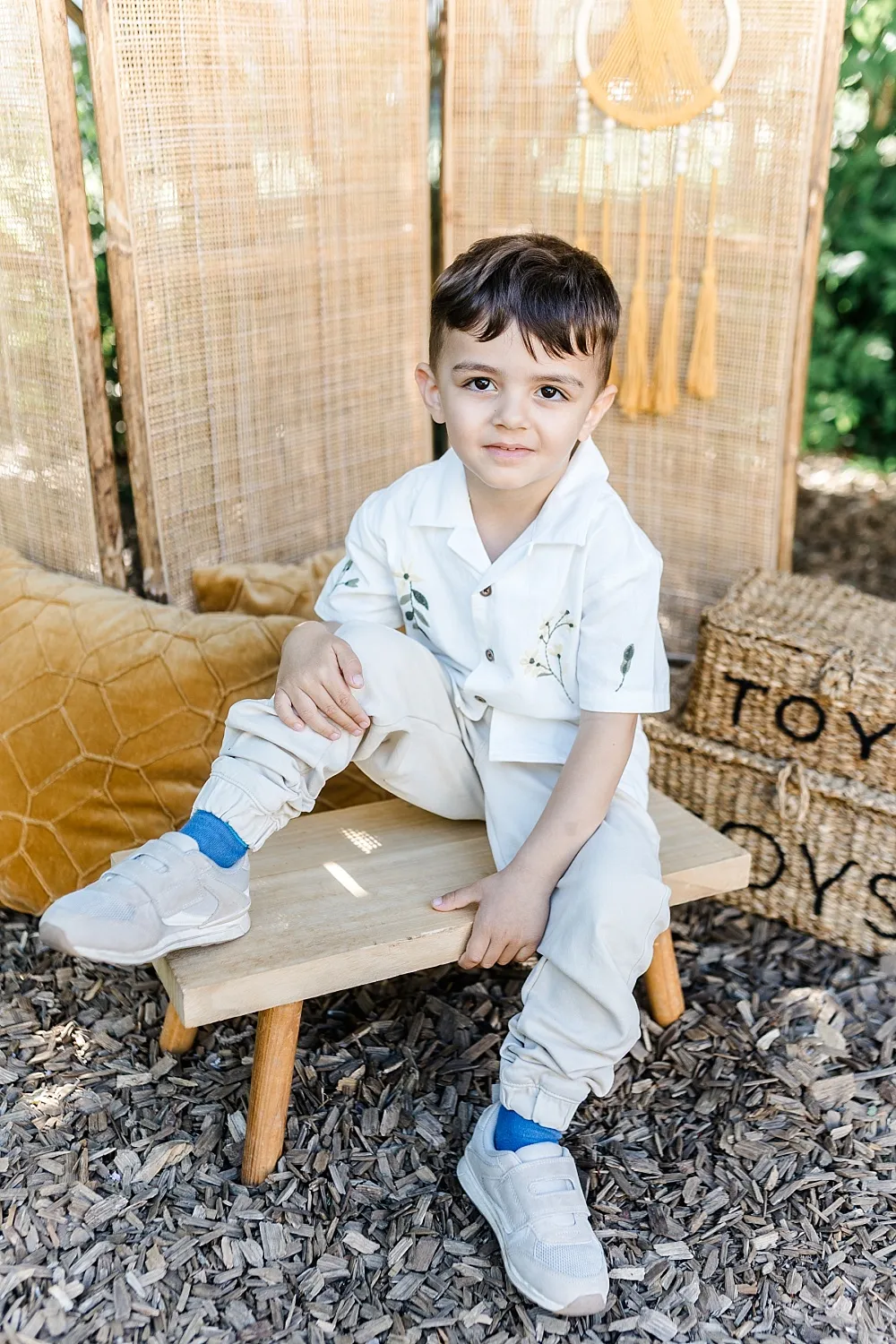 Kleiner Junge mit weißem Hemd und blauen Socken sitzt auf einer Holzbank im Außenbereich mit Spielzeugkörben im Hintergrund.