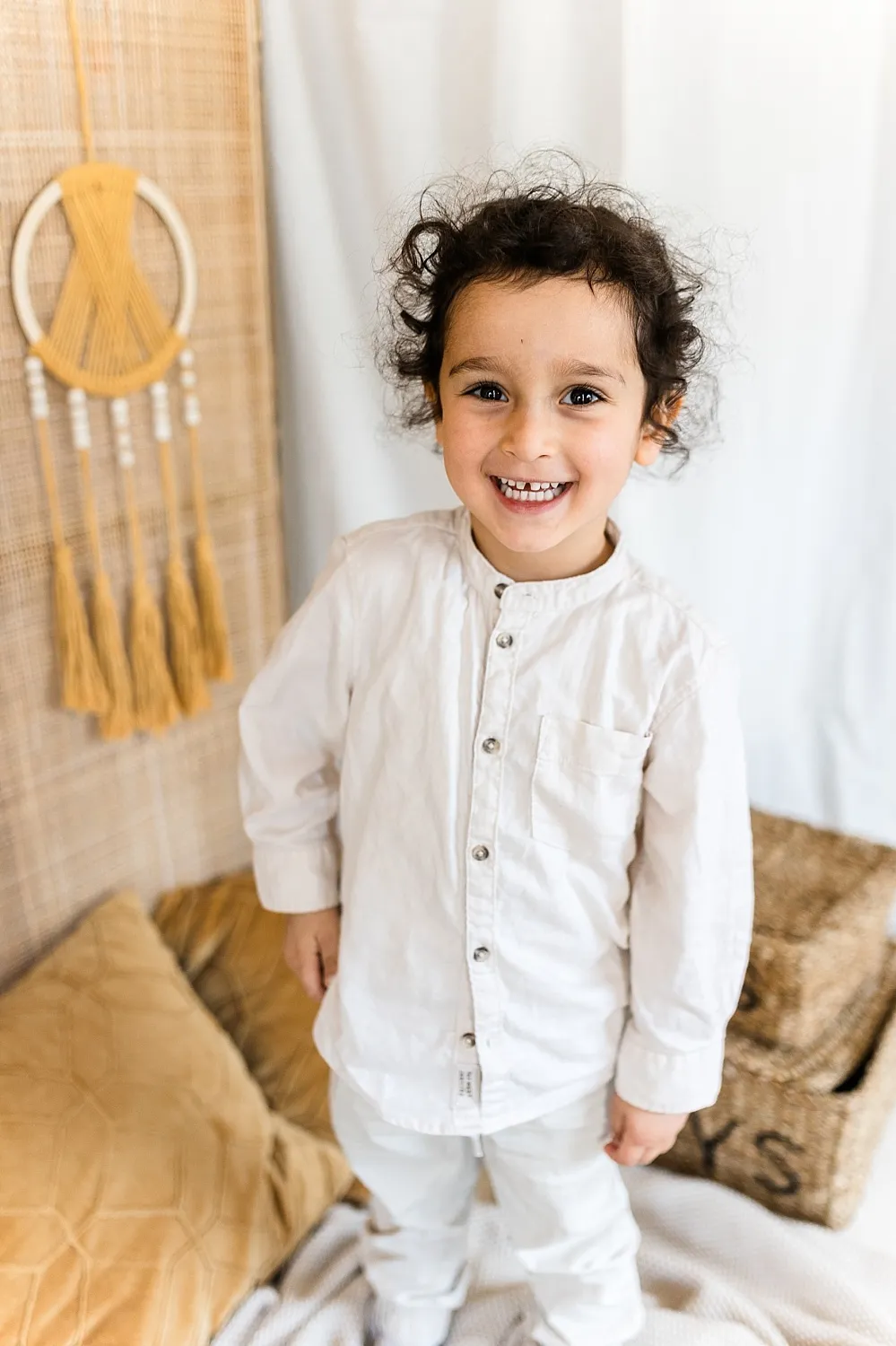 Fröhliches Kind mit lockigem Haar und weißem Hemd steht vor einem hellen Hintergrund mit dekorativen Körben und Kissen.