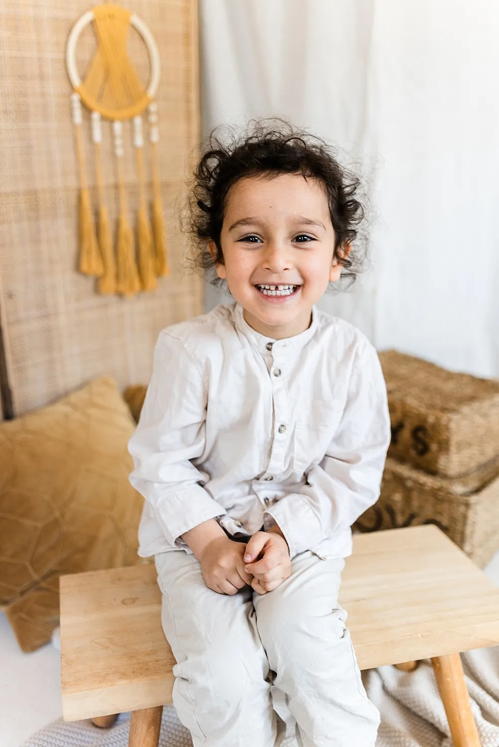 Lächendes Kind mit lockigem Haar und weißer Kleidung sitzt auf einer Holzbühne in einem gemütlichen Raum mit Teppich und Kissen.